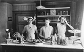 Tre expediter vid en väldfylld disk på ett hembageri i Varberg, omkring 1910. I mitten står ett fat med mandelmusslor, kringlor till vänster och olika småkakor i burkar och på glasfat. Flickan till vänster håller i en plåt med nybakade snittkakor och hon till höger visar att butiken är försedd med telefon. På hyllorna, dekorerade med hyllremsor, bakom dem ligger olika bröd och en stor burk med Mazettis cacao står i centrum. En skylt förkunnar att de är återförsäljare för Kanolds gräddkarameller. Både plåtask och vasen med rosor på disken visar upp jugendstilens estetik.
