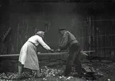 Johannes Lundgren och Berta Nilsson hjälps åt att hyvla bräder på Lilla Barkhult i Ry, Veddige. Hyveln har handtag på sidorna. Runt dem ligger spån och på väggen hänger en rubank.