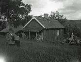 En man står på ängen och spelar på ett litet dragspel (durspel troligen) vid f d Torsorrens stuga. En kvinna sitter och hör på.