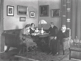 Borgmästare Johan H L Janssen med frun Annie, samt dottern Margareta med familj, som sitter i en soffgrupp. Väggen pryds av tavlor och till höger syns en stor jugendtextil med näckrosor och svanar. Bostaden ligger i ett av de s k 