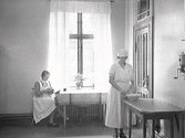 Tjänstefolk hos borgmästare Johan H L Janssen i köksregionen. En kvinna sitter vid fönstret och ser ut att putsa en kopparkittel medan den andra står med en bunke vid ett bord. På väggen sitter en kaffekvarn. Våningen låg i 