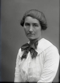 Kvinnoporträtt, sittande halvfigur. Kvinnan bär en stor rosett i halsen och lång halskedja på den vita blusen. Beställare: Hulda Bengtsson.