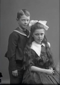 Syskonbild med en pojke med sjömanskrage och flicka i mörk klänning med stor vit snibbkrage och rosett i håret. Beställare: Tora Lambert.