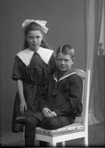 Syskonbild med pojke med sjömanskrage och flicka i mörk klänning med stor vit snibbkrage och rosett i håret. Beställare: Tora Lambert.
