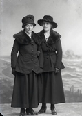 Två kvinnor i ytterkläder, där de lårlånga jackorna har stora pälskragar. Beställare: Elin Johansson.
