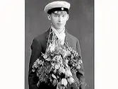 Examensporträtt av en yngling i studentmössa, Vidar Antonsson, Varberg. Vidar har blomsterbuketter hängande kring halsen och mössa med blank skärm.