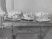 Tre detaljrika bakverk, kanske i kristyr, från Konditori Mignon uppställda på var sin bricka på ett bord. En föreställer en skottkärra och en annan en dragkärra.