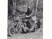 Militär i skogen på Albin-Monark motorcykel, 1953. Packväskor baktill. Han bär skinnhätta, mc-glasögon och har en väska över axeln.