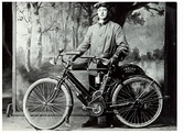 Ateljéfotografi av Anders Andersson från Stenstorp Gällared med första motorcykeln. De är avbildade mot en fond med skir lövskog vid ett vattendrag. Han medförde motorcykeln från USA 1909 då han var på besök i hemlandet. 