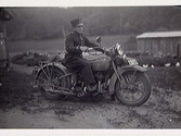 Evert Halltorp iklädd exerciskläder (militärkläder) på en motorcykel av märket Harley Davidson. Skärte 3, Rolfstorp. I bakgrunden höns och uthus.