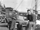 Näten lagas i Bua hamn, 1960-tal. En ung man lagar nät på kajkanten invid fiskebåten VG44. Bredvid står en liten pojke. Vid en trave fisklådor i bakgrunden står en motorcykel parkerad.