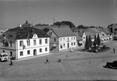 Bebyggelsen bakom Lagafontänen och dess fyra pelarträd på Stortorget i Laholm. Konstnären till fontänskulpturerna är John Lundqvist och fontänen invigdes 1933 av kung Gustav VI Adolf och drottning Louise. Vid Rådhusets gavel till vänster står en buss parkerad och några människor går över torget. I affärsbyggnaderna finns bland annat apotek, frisör och kemikalieaffär.