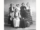 Sex unga kvinnor i olika folkdräktsliknande utstyrslar samt en avvikande som kan vara Frälsningsarméns dräkt med bahytt. Maskerad.