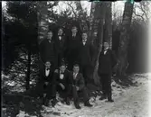 Åtta unga män i kostym och fluga står vid några björkar intill en väg i vintrig utomhusmiljö. Beställaren Erik Lundqvist är sannolikt med på bilden.