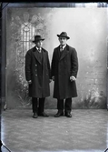 Ateljébild med två män i kostym, hatt och överrock, varav beställaren Gustav Johansson, Falkenberg sannolikt är en av dem. Troligen är bilden tagen i Björkströms Falkenbergsfilial.