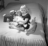 Familjen Arne Hedén, flicka sittandes i en säng tillsammans med sin docka, teddybjörn och leksakshäst