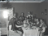 Elva kvinnor sitter vid ett bord varav ett par klädda i hucklen, hatt och/eller folkdräktslik utstyrsel. Man äter soppa, ost och bröd. Bilden är tagen i ett hem, Thorssons hus, med för tiden moderna, ljusa jugendmöbler. Beställare: Fröken Ljungberger.