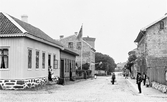 Till vänster närmast kameran syns kv Bagaren längs Prästgatan. Trädgården med flaggstång tillhör fotograf Mathilda Ranch. På huset till höger i kv Sparbanken hänger en skylt med texten: N. Göranson toffelmakare. Bilden är tagen mot väster så efter den korsande Kungsgatan ligger kv Magistern till vänster och kv Gästgivaren till höger.

Längs Prästgatan och även uppe i tornet centralt i bild står folk vända mot fotografen. Endast mannen i kubb till höger vänder sidan till.