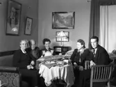 Gustaf Björkströms familj i vardagsrummet hos makarna Björkström i kv Smeden på Ringgatan 84, dit de flyttade 1940. Troligen firas makan Elins födelsedag då bordet är fullt av presenter och hon håller i en av gåvorna. Runt bordet sitter Elin med barnen, fr v Bo, Märta, Ulla och Åke. Över soffan hänger ett stort porträtt av Beethoven, kanske en av Gustafs musikaliska förebilder. (Se även bildnr GB2_6708, GB2_6709)