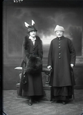Ateljébild av två kvinnor stående i helfigur i ytterkläder. Båda bär hatt; den ena pryds med fågelfjädrar likt vingar ovan kullen. Den yngre kvinnan har handväska, muff och stola. Den äldre håller ett paraply i ena handen och bär en kappa med stora, blanka knappar. Beställare: Emelie Nilsson.