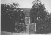 Författaren och läkaren August Bondesson står vid grinden till sitt barndomshem Fågelboet i Vessigebro, omkring sekelskiftet 1900. Intill honom står en blommande syrenbuske.