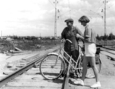 Småland hjälper kvinna med cykel vid övergång i Gustavsvik, 1950-tal