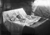 Dödsbäddsbild av ett dött barn nerbäddat i en säng. (Skadat negativ)