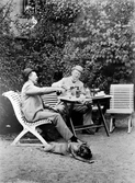 August Bondeson och vännen Hjalmar Carlsson sitter i en trädgårdsmöbel och slår i glasen ur buteljer. Båda bär kubb. På marken ligger en hund.