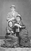 Från August Bondesons vänkrets. Ateljéfoto av två sittande kvinnor och en stående man, som döljer ena handen i rocken likt Napoleon. Anteckning på fotografiet baksida: 