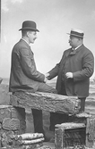 Atlejéfoto av två män som hälsar på varandra. Oscar Pehrson sitter på en rekvisitamur med björkved nedanför och mannen till höger heter Olle-Petter.