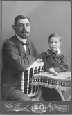 Handelsman Oscar Pehrson med sonen Håkan, född 1903. Oscar sitter bakvänd på en pinnstol och sonen står bredvid med armen på ett textilklätt bord, där det ligger en äggformad leksak.
