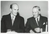 Två kostymklädda män sitter vid ett bord, 1950-tal. Till vänster ses Bengt Johansson (1930 - 2016) som ägde gården Labacka Lund. Mannen till höger som håller en cigarett är okänd.