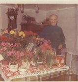 80-årsuppvaktning för David Bengtsson (1889 - 1977), Långåker 1:2 1969. David står snett bakom ett bord som är fyllt av blommor. Ett väggur skymtas i bakgrunden.
