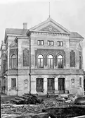 Ur grosshandlare Oskar Nordbloms fotoalbum. Entréfasaden av Varbergs teater vid Engelbrektsgatan, där vägarbete pågår. Timmerstockar och stora stenstycken ligger framför teatern som uppfördes 1895.