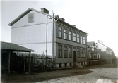 Ur byggmästare Johannes Nilssons fotoalbum från 1914. Bebyggelse utmed Eskilsgatan i kv Oxen, Varberg, med gatunummer 36 i förgrunden. Vid husgaveln står en elektrifierad gatubelysning, direkt kopplad till elledningen som korsar gatan.