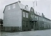 Ur byggmästare Johannes Nilssons fotoalbum från 1914. Flerbostadshus uppfört 1914 i två våningar på Slottsgatan 12 i kv Matrosen, sammanbyggt med grannhuset. Plåttaket ser skinande nytt ut och har en något ovanlig utformning: plant brutet med branta takfall (se även byggmästarens hus på Slottsgatan 15 bildnr VMA11716 D33). Det medger största möjliga rumsyta med god takhöjd även på vindsvåningen, som förses med ljus från  runda takfönster (lukarner) och två frontespiser. Över entrén sitter en balkong med elektrisk fasadbelysing, trots att lyktstolpen står precis intill. Intill flaggstångsfästet angör elledningarna huset.
