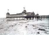 Ur byggmästare Johannes Nilssons fotoalbum från 1914. Kallbadhuset i Varberg som uppfördes 1904. De vita skynkena under badhuset skyddar de badande  från insyn. På stranden leker barn i olika åldrar.