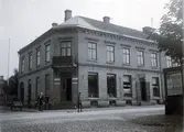 Ur byggmästare Johannes Nilssons fotoalbum från 1914. Byggnaden i korsningen Kungsgatan-Prästgatan, kv Bagaren och lär vara uppfört på 1860-talet. På hörnet ligger entrén till Victor Perssons bageri och konditori. Vidare längs Kungsgatan ligger butiken Grand Bazar. Runt hörnet in på Prästgatan står en tom kärra. På plankhörnet sitter affischer från Kindvalls Tryckeri som annonserar föreställningen Pygmalion med Harriet Bosse på Teatern. Dramaten gjorde under 1914 detta gästspel, vilket ger fotografiet sin datering.