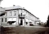 Ur byggmästare Johannes Nilssons fotoalbum från 1914. Vy över fastigheten där Oscar Pehrsons speceriaffär, OP:s kallat, låg i korsningen av Kungsgatan-Norrgatan, kv Lagmannen. På skyltfönstren görs reklam för bland annat ångrostat kaffe, viner och konserver. I samma byggnad låg vid Norrgatan två skrädderier, både systerns Augusta Pehrsons och Jean Svenssons.