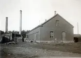 Ur byggmästare Johannes Nilssons fotoalbum från 1914. Okänd tegelbyggnad i Falkenberg. Möjligen ett sågverk då olika typer av virke ligger utanför byggnaden. Anläggningen har en rund och en fyrkantig skorsten. Meddela oss gärna om du har information kring anläggningen!