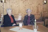 Två kvinnor sitter bakom ett bord i Missionskyrkan, okänt årtal. Från vänster: Aslaug Benkel (1925 - 2018) och Maja Gunnarsson (1919 - 2015). Väggarna i bakgrunden är klädda med träpanel.