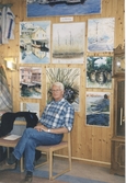 Hantverksutställning i Missionskyrkan, okänt årtal. Tor Söderberg sitter på en stol framför en vägg fylld av tavlor. Väggarna är klädda med träpanel.