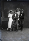 Ateljéfoto av två kvinnor och två män i helfigur framför en stor tavla där männen sitter på en balustrad. Samtliga är iförda hattar. Beställare: Anna Reinholdsson.