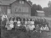 Skolfoto taget utanför en skola på landsbygden. Lärarinnan står till höger och barnen ser ut att ha fått instruktion om att lägga armarna i kors. Meddela oss gärna om du vet var skolan ligger!