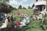 Kållereds Missionsförsamlings barnkör har våravslutning år 2000 i trädgården hemma hos Rune och Ann-Marie Karlsson, Labacka. Ann-Marie (vithårig) ses gående till höger vid bostadshuset.