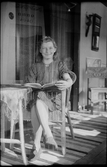 Ella Lundgren sitter och läser i en korgstol. Hon bär klänning och skor i matchande mönster och en halsbrosch. Bilden är dubbelexponerad med ett foto taget vid den öppna bemålade spisen i hennes hem, där en pojke sitter.