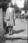 Ella Lundgren står i rutig kjol och jacka vid en högsmal sten och bikupor vid granarna bakgrunden.