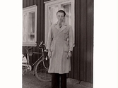 En ung man klädd i överrock, Karl Hugo Svensson, fotograferad utanför Vänhem (Gerda Winqvists bostad). Karl Hugo arbetade i Rosenqvists affär (Eftr). En cykel står vid husväggen bakom honom.