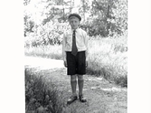 Gossen Karl Hugo Svensson, klädd i basker, skjorta med slips och kortbyxor, fotograferad utomhus i en trädgård. Han var fosterson hos Augusta Karlsson i Tvååker.