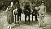 Karl och Alice Snygg står utomhus med tre hästar emellan sig. Karl är arbetsklädd men Alice bär klänning och spetsiga skor med rem.